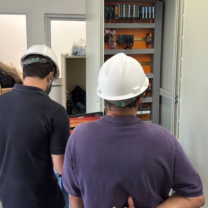 Duas pessoas de costas trabalhando com notebook diante de um painel elétrico aberto.